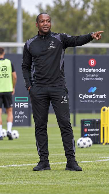 Lennux Ferdinand coaching on a football pitch with technology partner boards visible in the background